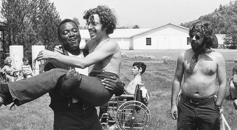 Outdoors in the summer. Three shirtless smiling men in the foreground. One is carrying the other. In the background a young man in a wheelchair in the grass.